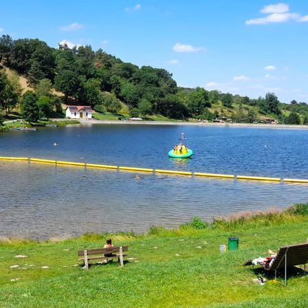 Eine ruhige Seelandschaft mit einem kleinen Häuschen, Menschen, die eine Flöße genießen, und anderen, die schwimmen. Gelbe Schwimmhilfen säumen den Wasserrand.