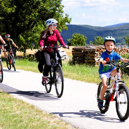 Eine Gruppe von Menschen fährt auf einem Betonweg mit Fahrrädern. Eine Frau und ein Kind sind vorne. Sie sind von Gras und einem Holzstapel umgeben. Berge sind in der Ferne.
