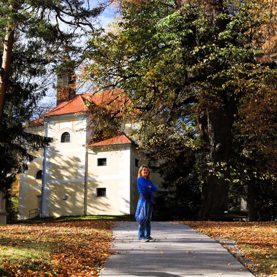 Eine Frau in einem blauen Outfit steht vor einem alten Gebäude mit einem roten Dach. Der Weg ist von Bäumen und fallenden Blättern umgeben.
