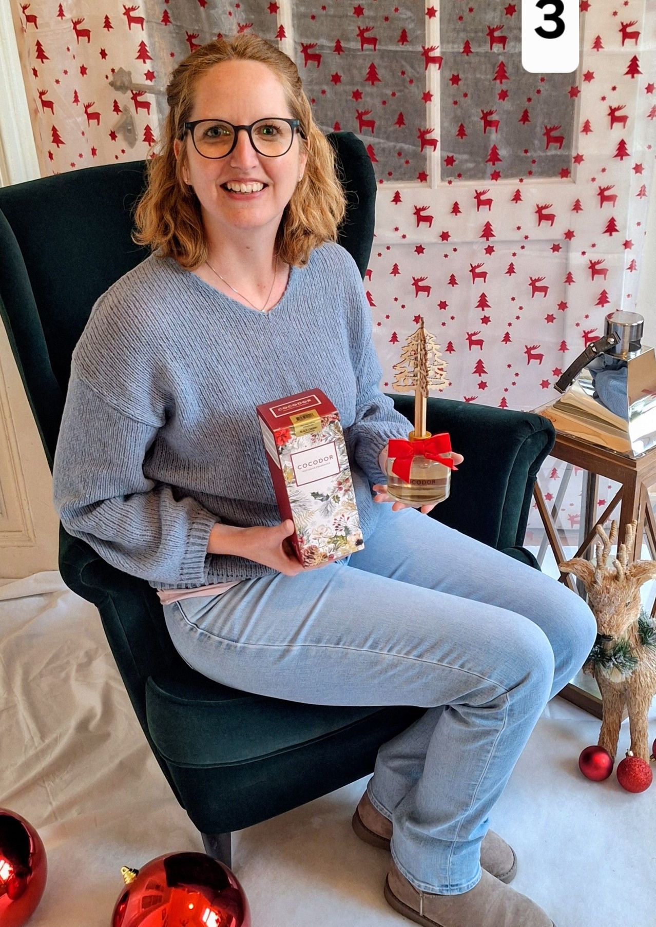 Eine lächelnde Frau in einem blauen Pullover hält eine rote Box und eine Weihnachtsbaumdekoration, während sie auf einem Stuhl vor einem festlichen Hintergrund sitzt.