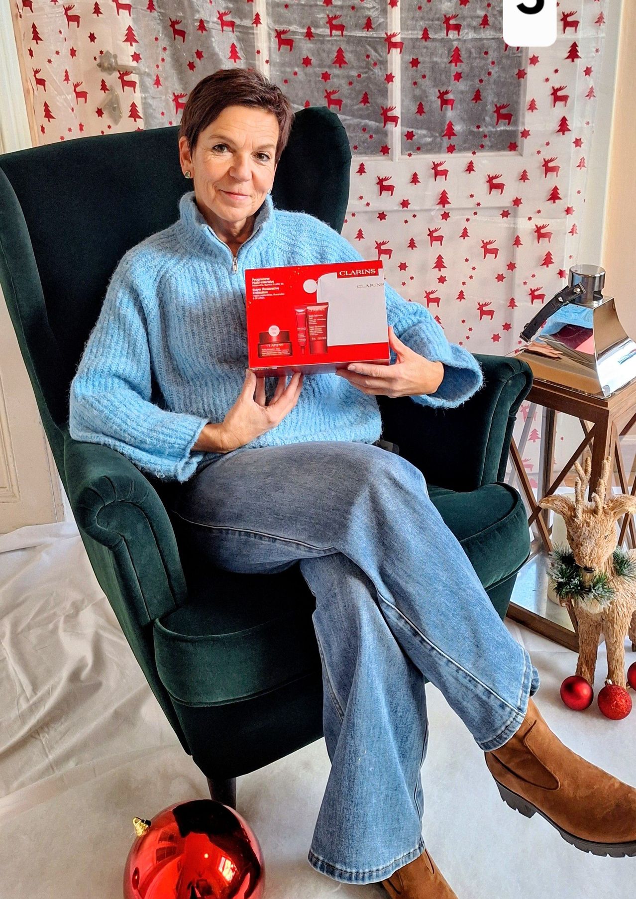 Eine Frau in einem gemütlichen blauen Pullover und Jeans sitzt auf einem grünen Sessel und hält eine rote Box mit dem Wort 'Clarins'. Hinter ihr befindet sich eine weihnachtlich gestaltete Wand mit roten Sternen und Rentieren.