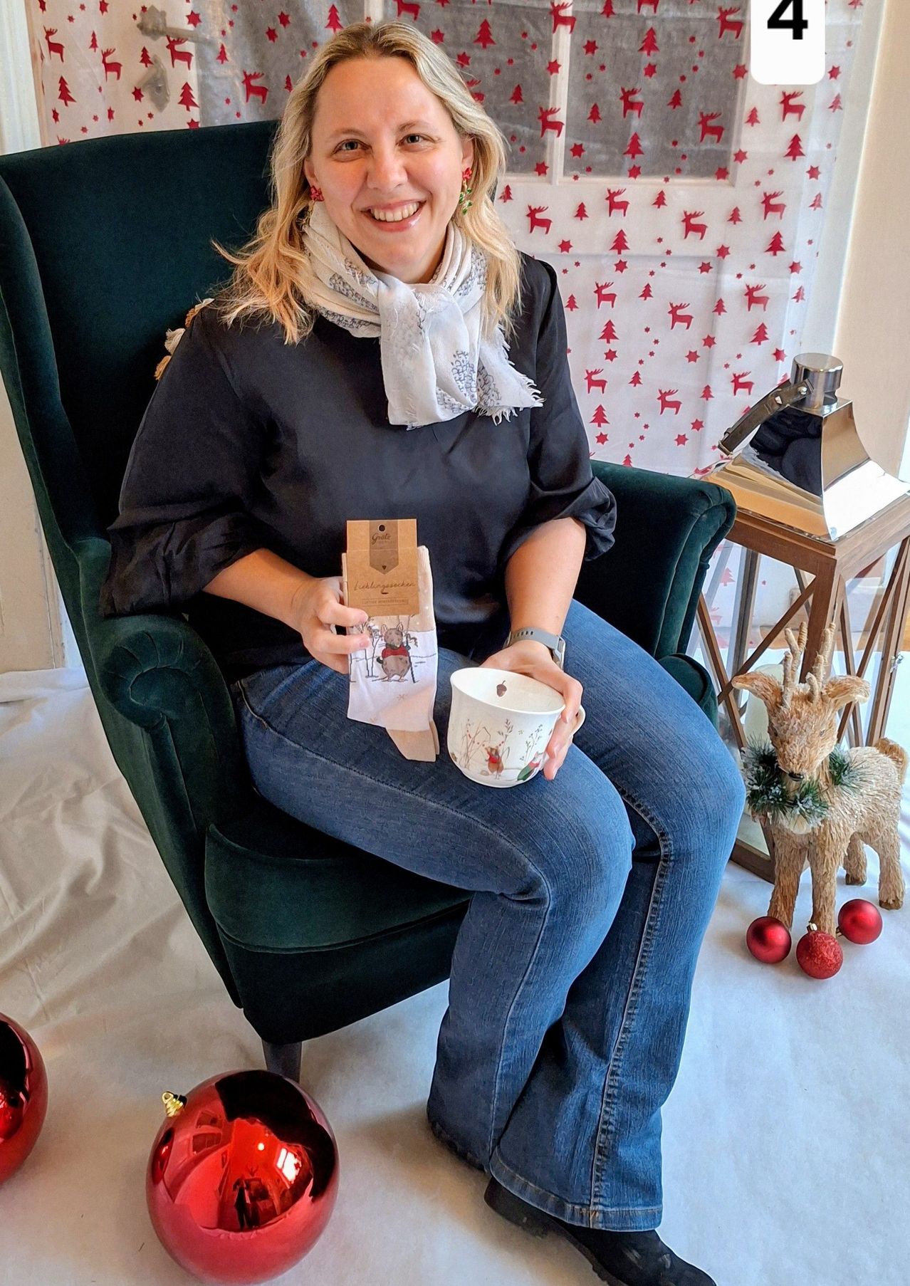 Eine Frau sitzt auf einem grünen Sessel, lächelt und hält eine Tasse und ein Paket. Sie trägt ein schwarzes Oberteil und Jeans. Hinter ihr ist ein Weihnachtshintergrund mit roten Sternen und Rentieren. Eine Rentierfigur und Weihnachtskugeln liegen auf dem Boden.