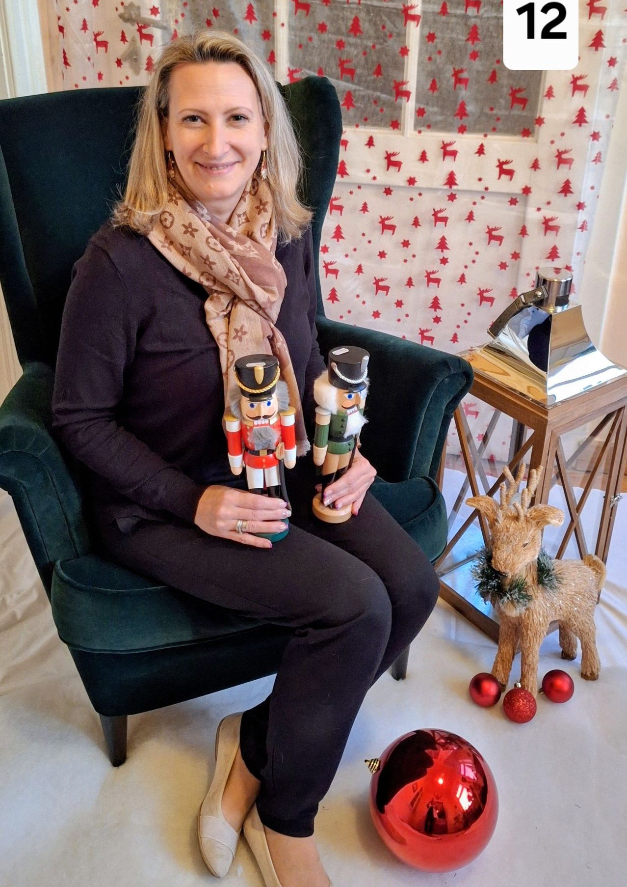 Eine Frau sitzt auf einem grünen Stuhl, hält zwei Nussknacker im Schoß, mit einem Rentier und Ornamenten in der Nähe. Hinter ihr ist ein weihnachtlicher Hintergrund.