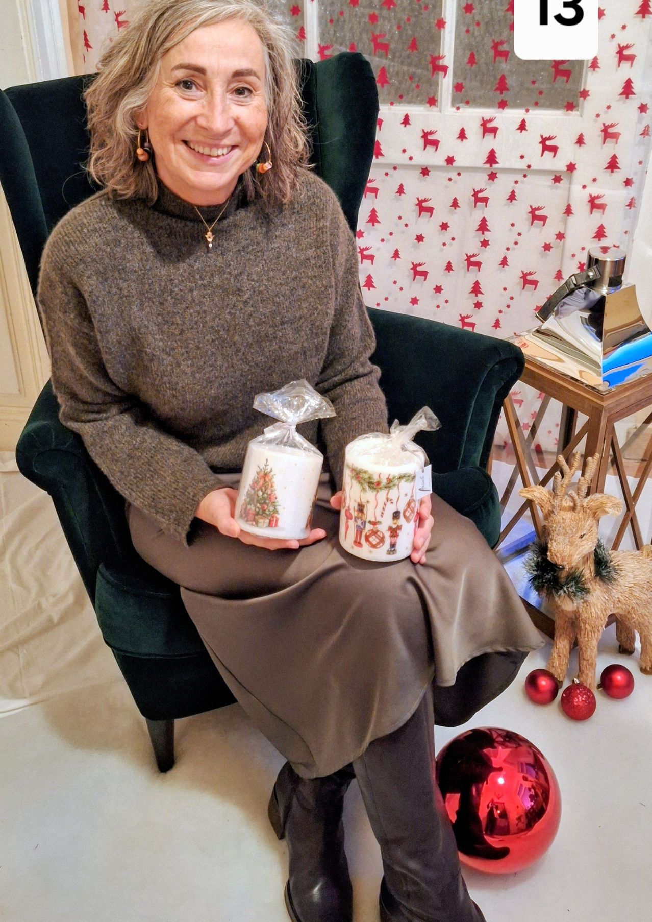 Eine Frau sitzt auf einem grünen Stuhl und hält zwei verpackte Weihnachtskerzen. Sie lächelt und scheint fröhlich. Hinter ihr ist ein festlicher Hintergrund mit Rentieren und Weihnachtsbäumen zu sehen. Auf dem Boden liegen auch Weihnachtsdekorationen.