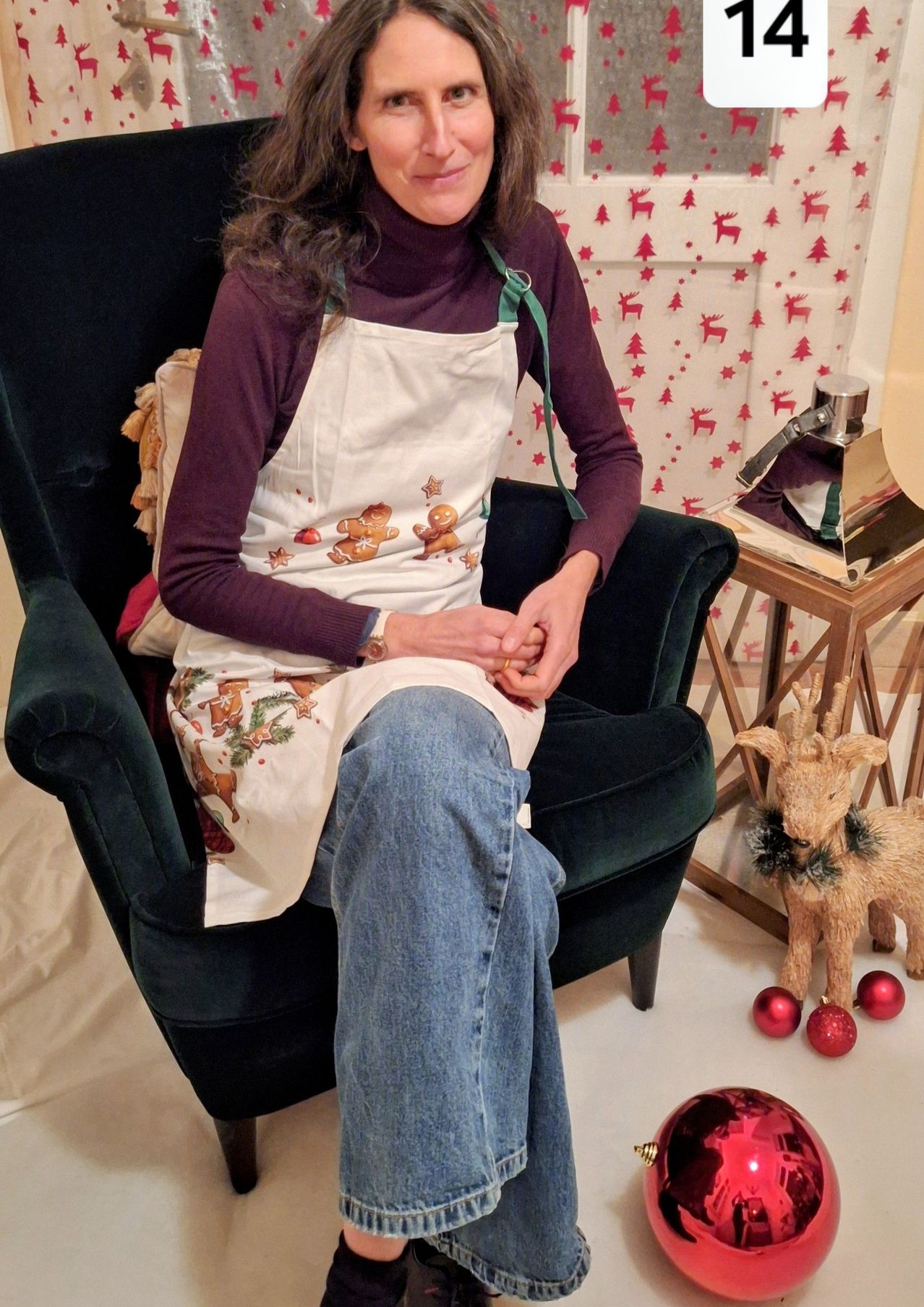 Eine Frau sitzt auf einem grünen Sessel und trägt eine Weihnachtsschürze mit Lebkuchen. Sie hält einen Keks in den Händen. Ein Weihnachtsbaum und Dekorationen sind im Hintergrund.