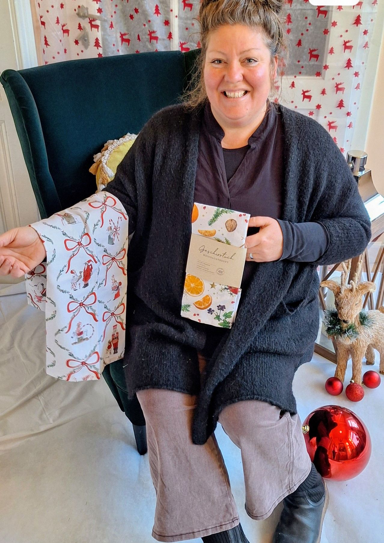 Eine Frau sitzt in einem Stuhl und hält einen festlichen Geschenkbeutel mit einem Design aus Orangen und Grün. Sie lächelt für die Kamera, mit einem Weihnachtsbaum und Ornamenten im Hintergrund.