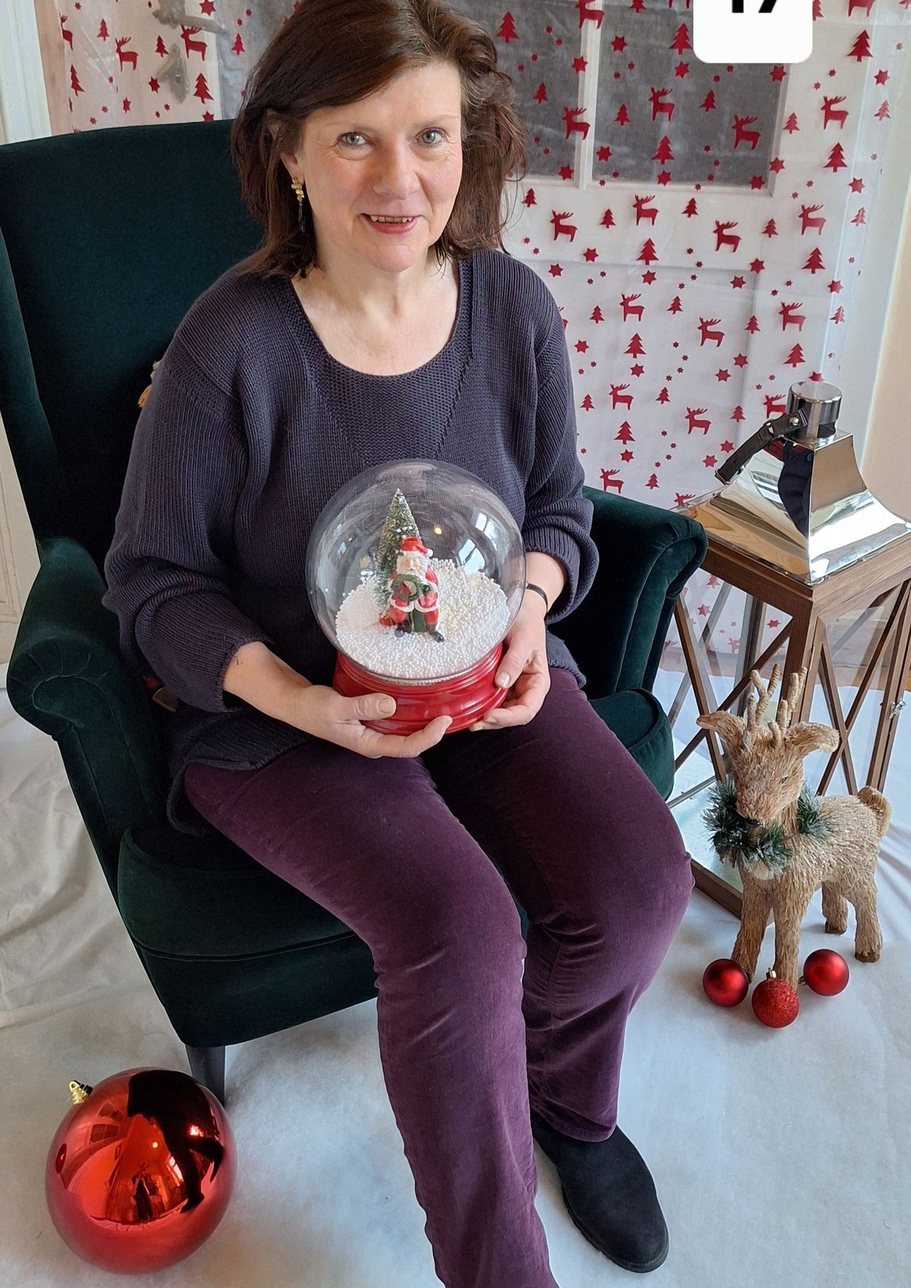 Eine Frau sitzt in einem Stuhl und hält eine Schneekugel mit Santa darin, umgeben von Weihnachtsdekorationen.