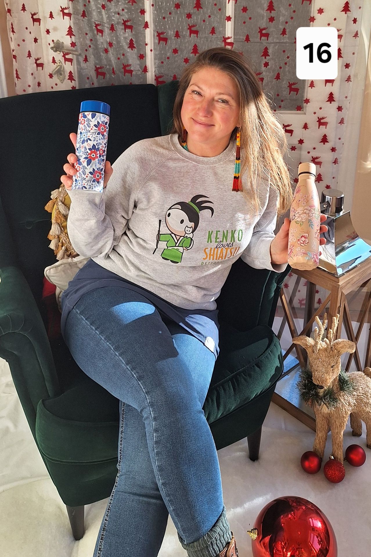 Eine Frau, die ein graues Sweatshirt mit einem Cartoon-Charakter trägt, sitzt auf einem grünen Stuhl und hält zwei Thermoskannen in den Händen. Hinter ihr steht ein Holztisch mit einer Rentierfigur und ein Weihnachtsbaum mit roten Sternen an der Wand.