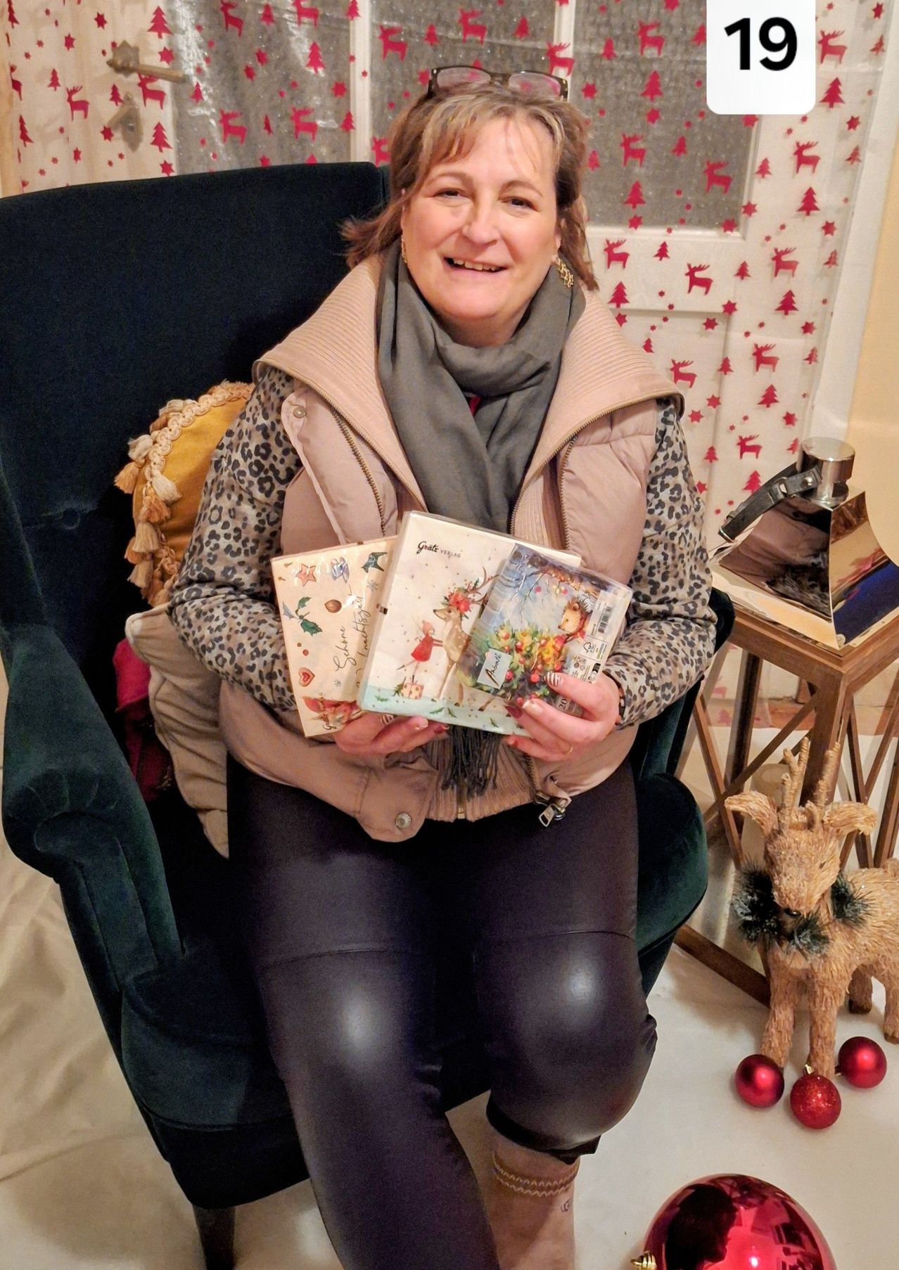 Eine Frau sitzt auf einem Stuhl, lächelt und hält Weihnachtskarten in den Händen. Der Hintergrund zeigt Weihnachtsdekorationen, darunter Rehe und einen Weihnachtsbaum.