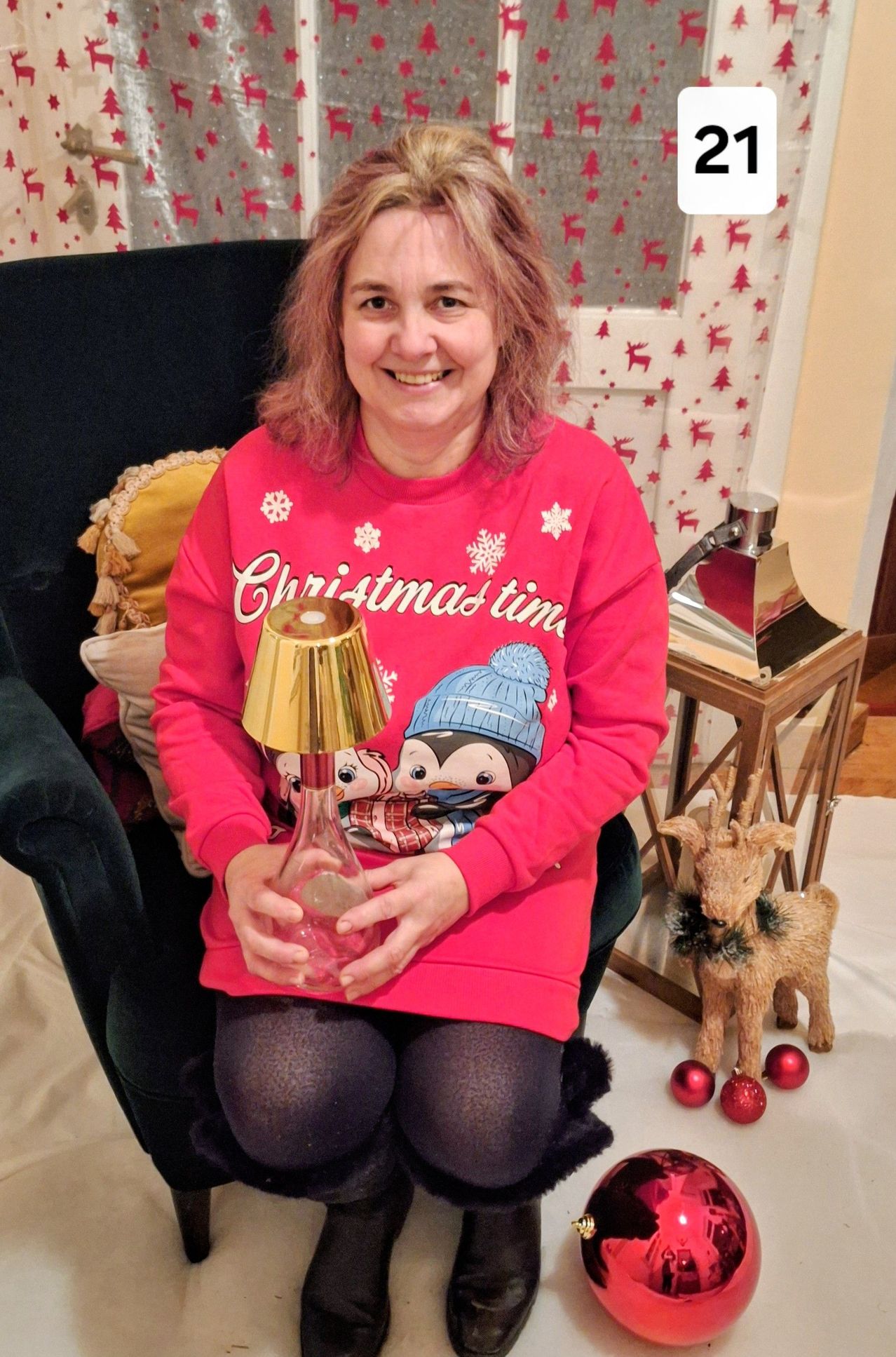 Eine lächelnde Frau in einem Weihnachtspullover posiert mit einem goldfarbenen Lampenschirm in einer festlichen Umgebung.