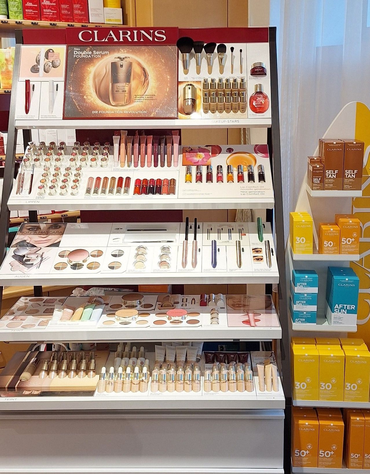 A display of various cosmetic products on shelves, including foundation, eyeshadow, lipstick, and other makeup essentials. A large poster in the center advertises a foundation revolution.