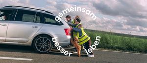 Drei Menschen in Warnwesten schieben ein silbernes Auto am Straßenrand, begleitet von einem braunen Hund. Die Worte 'Gemeinsam besser leben' sind prominent angezeigt.