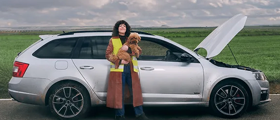 A woman in a reflective vest stands beside a silver car with its hood open, holding a small brown dog on a grassy road.