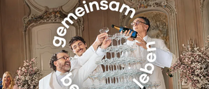 Drei Männer in weißen Anzügen gießen Champagner in eine Pyramide aus Gläsern auf einem Tisch, mit einer Frau in Pink, die hinter ihnen steht.