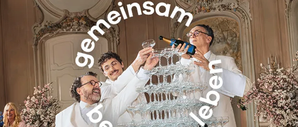 Drei Männer in weißen Anzügen gießen Champagner in eine Pyramide aus Gläsern auf einem Tisch, mit einer Frau in Pink, die hinter ihnen steht.