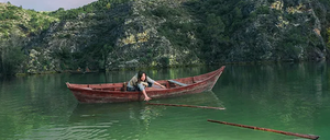 Eine Frau paddelt ein kleines rotes Boot auf einem See mit einem felsigen Ufer und üppiger Vegetation im Hintergrund.
