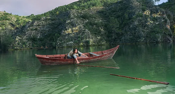 Eine Frau paddelt ein kleines rotes Boot auf einem See mit einem felsigen Ufer und üppiger Vegetation im Hintergrund.