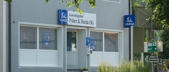 Ein Bürogebäude mit einem Schild, das GeneralAgentur Priber & Durda OG besagt. Das Gebäude hat weiße Fenster und eine Tür. Es gibt ein Schild mit einem grünen Pfeil, der nach rechts zeigt.