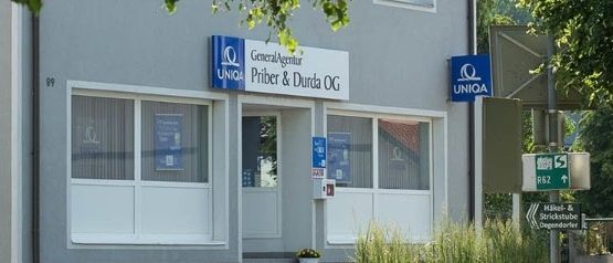Ein Bürogebäude mit einem Schild, das GeneralAgentur Priber & Durda OG besagt. Das Gebäude hat weiße Fenster und eine Tür. Es gibt ein Schild mit einem grünen Pfeil, der nach rechts zeigt.