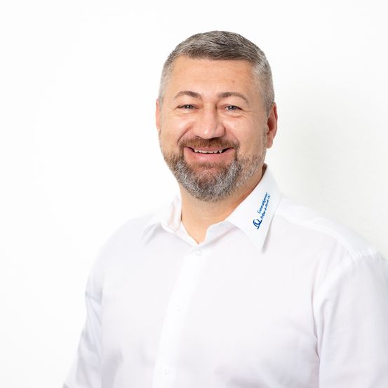 Ein lächelnder Mann in einem weißen Hemd mit einem Logo auf der linken Brustseite steht vor einem weißen Hintergrund.