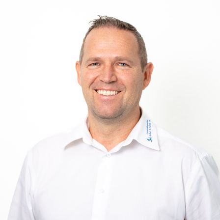 Ein Mann in einem weißen Hemd mit einem Logo auf der linken Brust, lächelt in die Kamera. Er hat kurze Haare und einen glattrasierten Gesichtsbart.