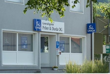 Ein graues Gebäude mit mehreren Fenstern und einer Tür. Es hat ein Schild, das GeneralAgentur Priber & Durda OG und ein Uniqa-Logo zeigt. Ein Topf mit Pflanzen steht vor der Tür.