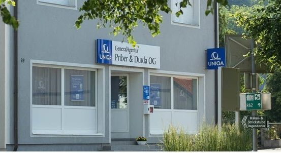 Ein graues Gebäude mit mehreren Fenstern und einer Tür. Es hat ein Schild, das GeneralAgentur Priber & Durda OG und ein Uniqa-Logo zeigt. Ein Topf mit Pflanzen steht vor der Tür.