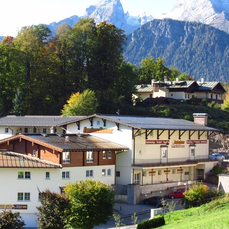 Ein weißes Gebäude mit braunen Dächern und mehreren Fenstern befindet sich in einem grünen Tal mit Bergen im Hintergrund. Bäume umgeben das Gebiet, und ein rotes Auto ist vor dem Gebäude geparkt.