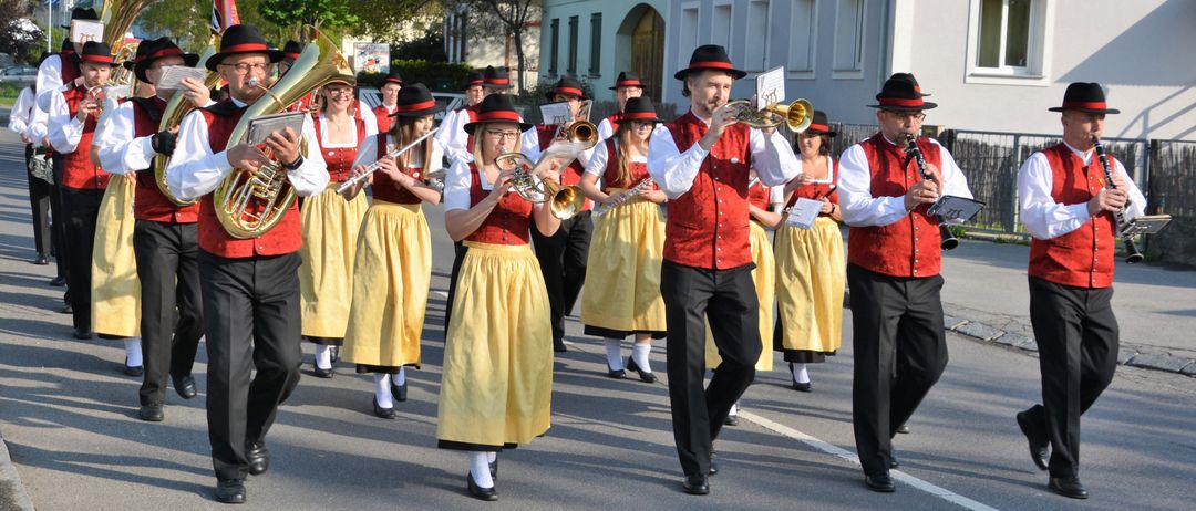 Eine Blaskapelle in rot und gelb gekleidet spielt Blechblasinstrumente und marschiert vor einem Haus die Straße hinunter.