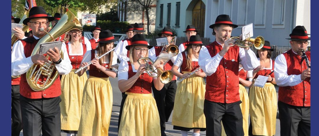 A marching band is performing on the street. They are wearing red vests and yellow skirts. The text on the image reads 'Maimarsch am 1. Mai'.