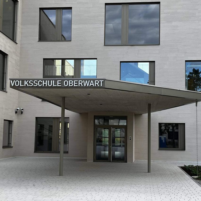 Ein modernes Gebäude mit Glasfenstern und Türen, namens Volksschule Oberwart, hat einen Vorbau mit einem Schild über dem Eingang.