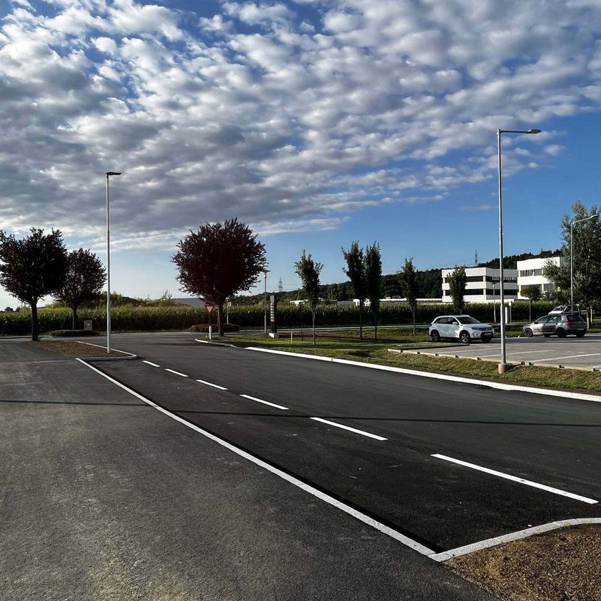 Eine leere Straße mit weißen Linien, Bäumen und Straßenlaternen. Ein weißes Auto ist auf dem Parkplatz rechts geparkt. Ein Gebäude ist in der Ferne.
