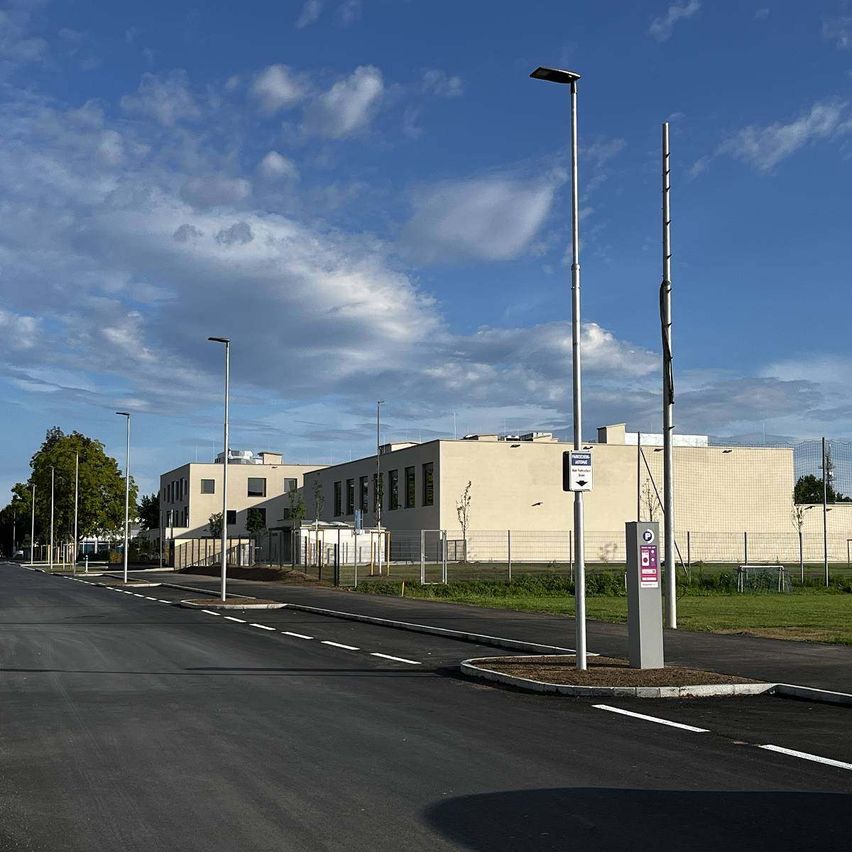 Eine leere Straße mit Straßenlaternen und einem Gebäude im Hintergrund. Die Straße hat weiße Markierungen, und es gibt zwei Masten mit Schildern. Der Himmel ist blau mit einigen Wolken.