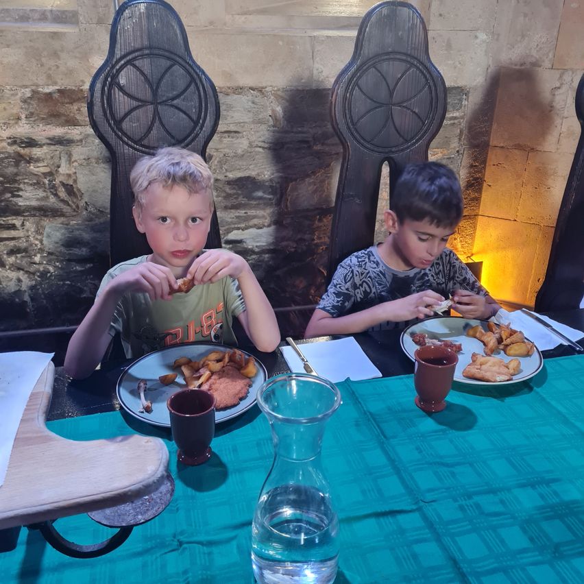 Zwei Jungen sitzen an einem mittelalterlichen Tisch und genießen ein Essen. Jeder hat einen Teller mit Essen und einen kleinen Becher. Der Tisch ist mit einem türkisfarbenen Tuch bedeckt.