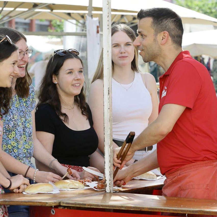 Ein Mann in einem roten T-Shirt grillt Essen an einem Tisch, während vier Frauen zuschauen und sich mit ihm unterhalten.