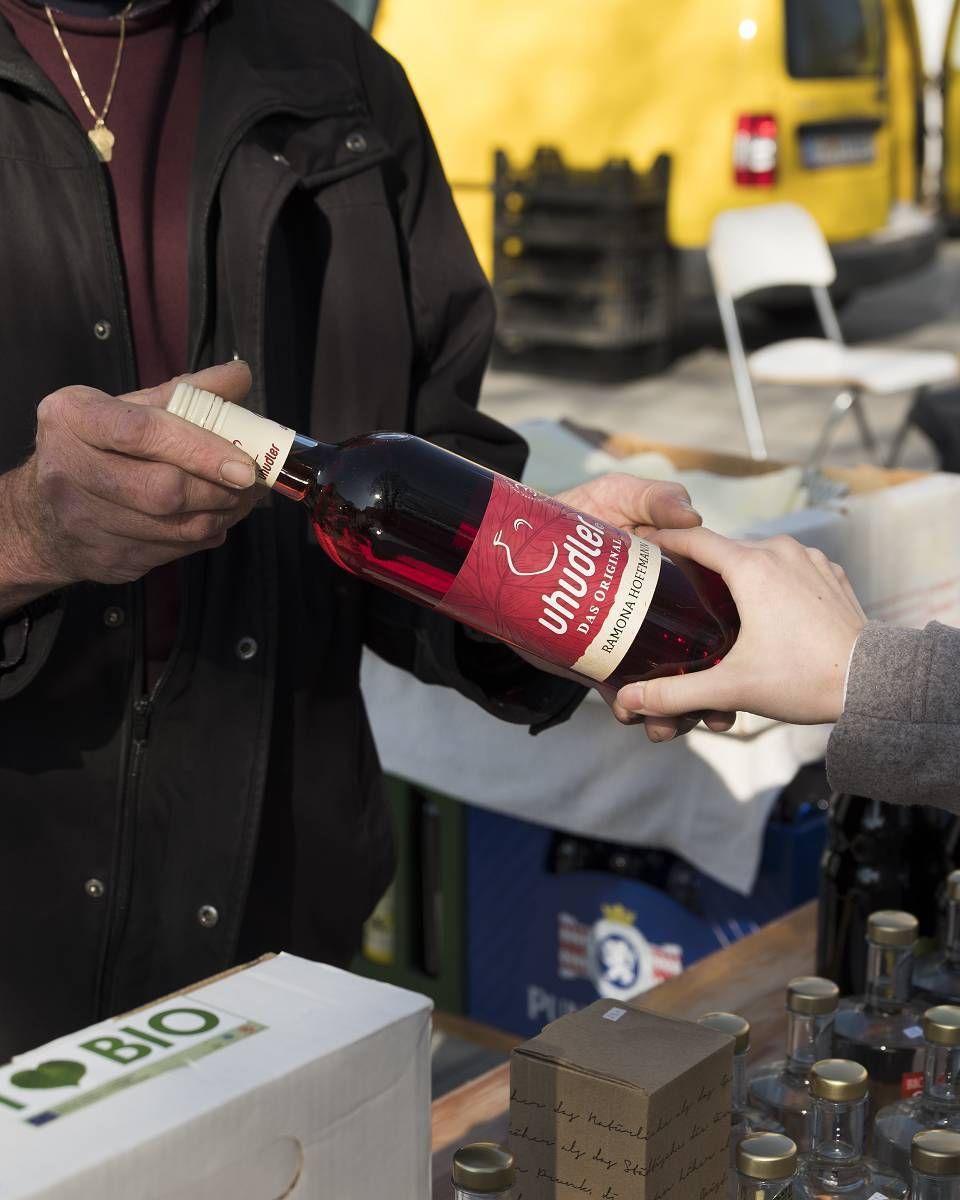 Eine Person in einer schwarzen Jacke gießt eine Flasche roten Weins in die Hand einer anderen Person auf einem Markt. Ein gelber Van und ein weißer Klappstuhl sind im Hintergrund.
