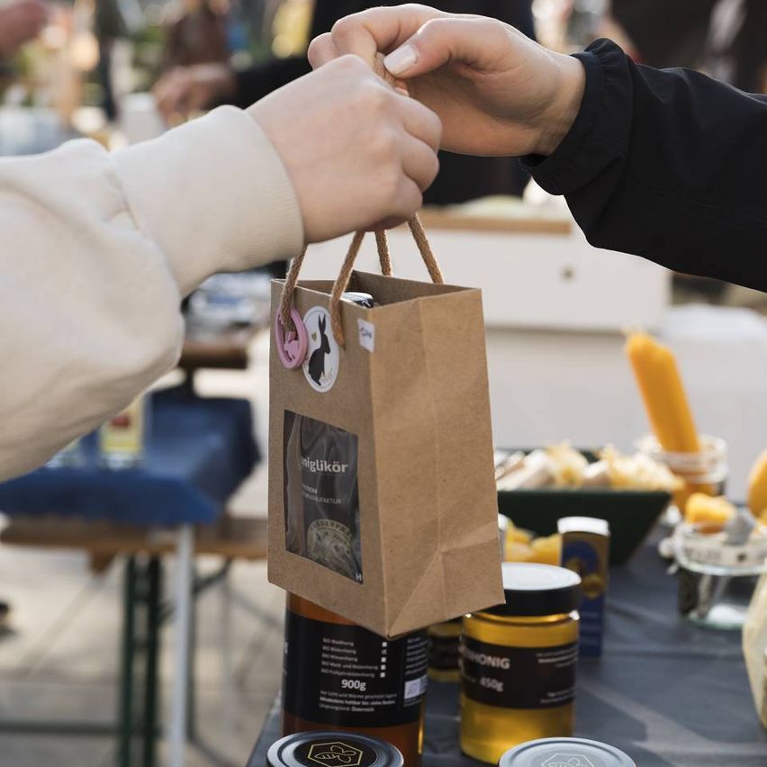 Eine Person, die einen braunen Papierbeutel an eine andere Person auf einem Markt übergibt. Der Beutel hat ein Etikett mit der Aufschrift 'niglikor'. Verschiedene Gläser und Behälter stehen auf dem Tisch.