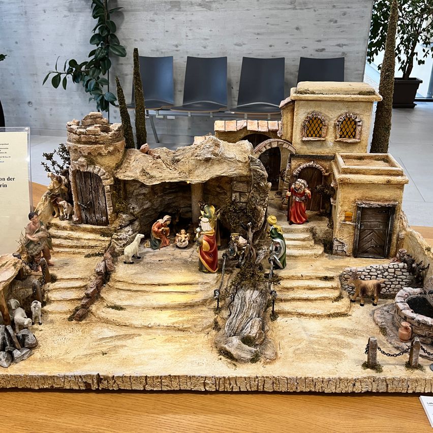 Eine kleinformatige Krippe aus Ton und Stein steht auf einem Holztisch und wird von einer Kulisse eines kleinen Dorfes und Grünpflanzen umgeben.