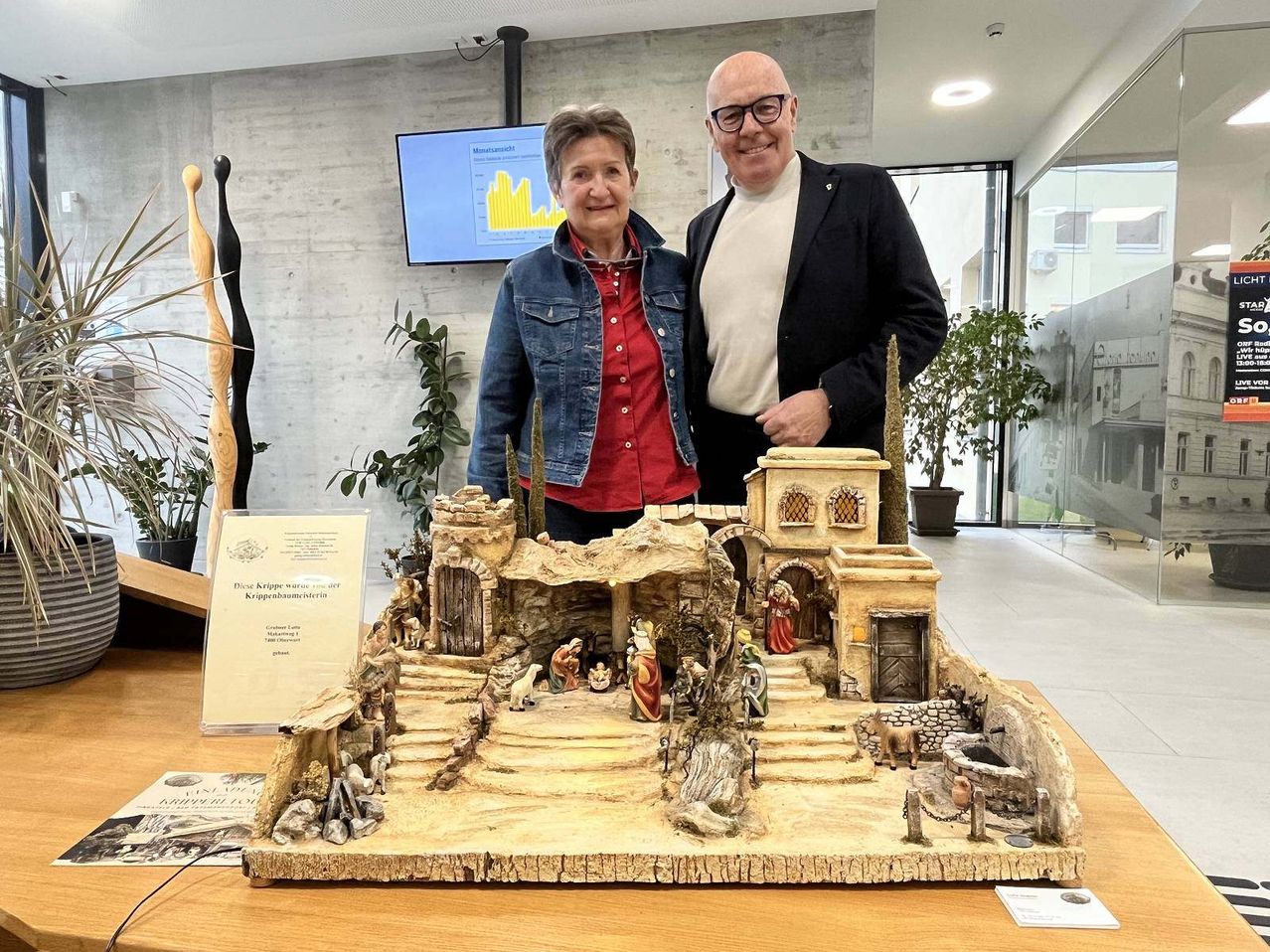 Zwei Personen stehen lächelnd hinter einer hölzernen Krippe. Die Szene zeigt eine Futterkrippe, Gebäude und Figuren von Menschen und Tieren. Ein Monitor ist an der Wand montiert, und Pflanzen sind im Raum platziert.