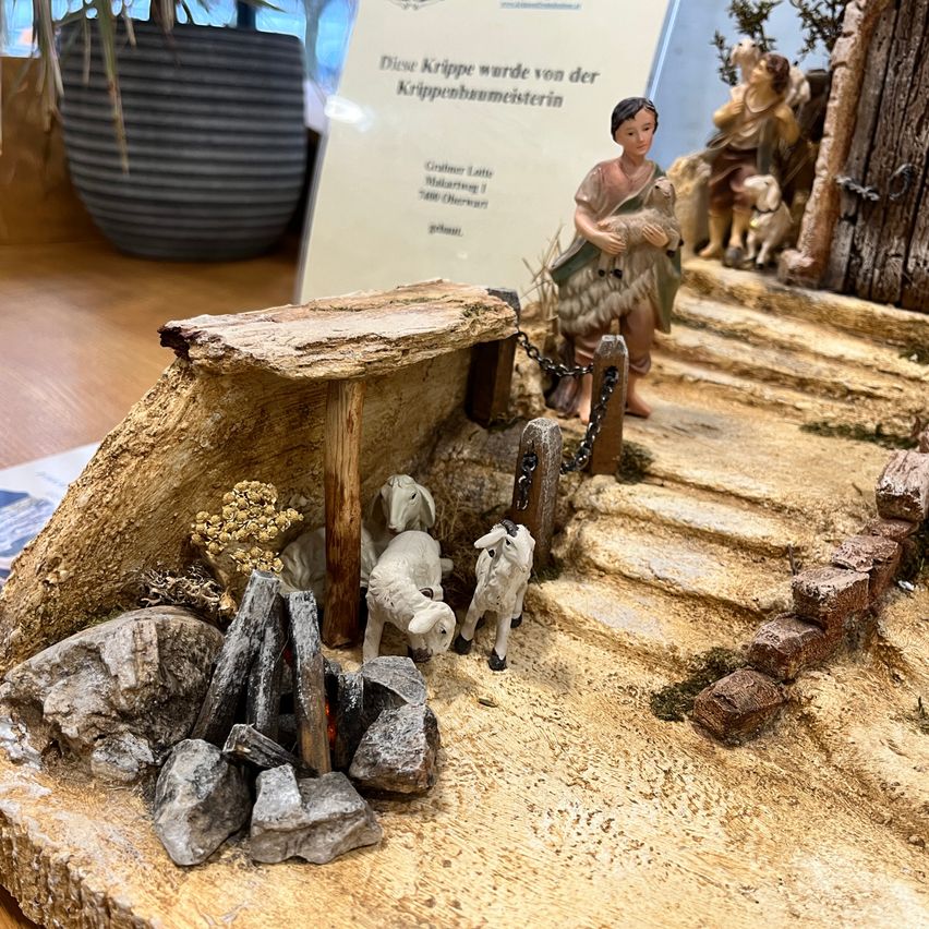 Eine Miniaturkrippe wird mit einem Hirten, der ein Lamm hält, einem kleinen Holzhaus und einem kleinen Feuerstellen mit einem Haufen Steinen und Zweigen ausgestellt.