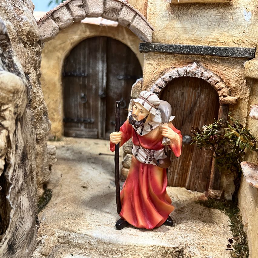 Eine Figur eines älteren Mannes mit Bart, der einen Umhang trägt und einen Stab hält, steht neben einer steinernen Bogentür in einer kleinen, rustikalen Szene.