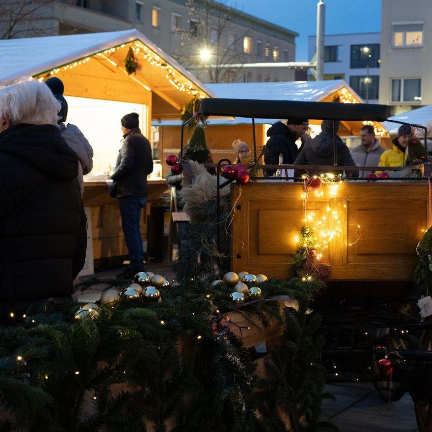 Ein Weihnachtsmarkt mit Holzhütten, beleuchtet mit Weihnachtslichtern, Menschen stöbern durch Stände und eine Pferdekutsche ist geschmückt.