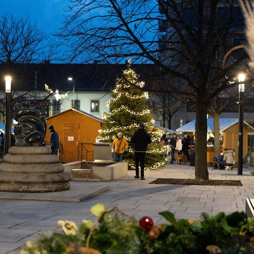 Bei Nacht schlendern Menschen durch einen Weihnachtsmarkt. Eine geschmückte Weihnachtsbaum steht in der Mitte, mit kleinen Geschäften auf beiden Seiten. Eine Statue steht auf einem Podest links.