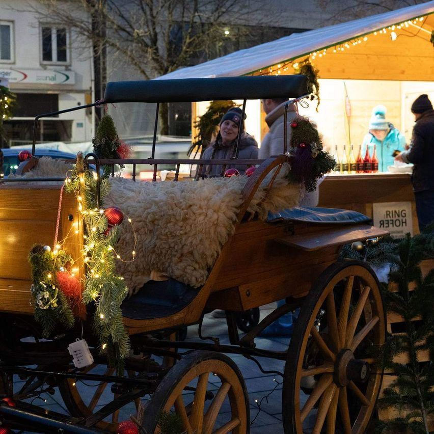 Ein holzernen Wagen mit einem mit Wolle bedeckten Sitz ist mit Weihnachtslichtern und Ornamenten dekoriert. Eine Frau mit Mütze steht dahinter und eine Person steht neben einem Stand mit Flaschen.
