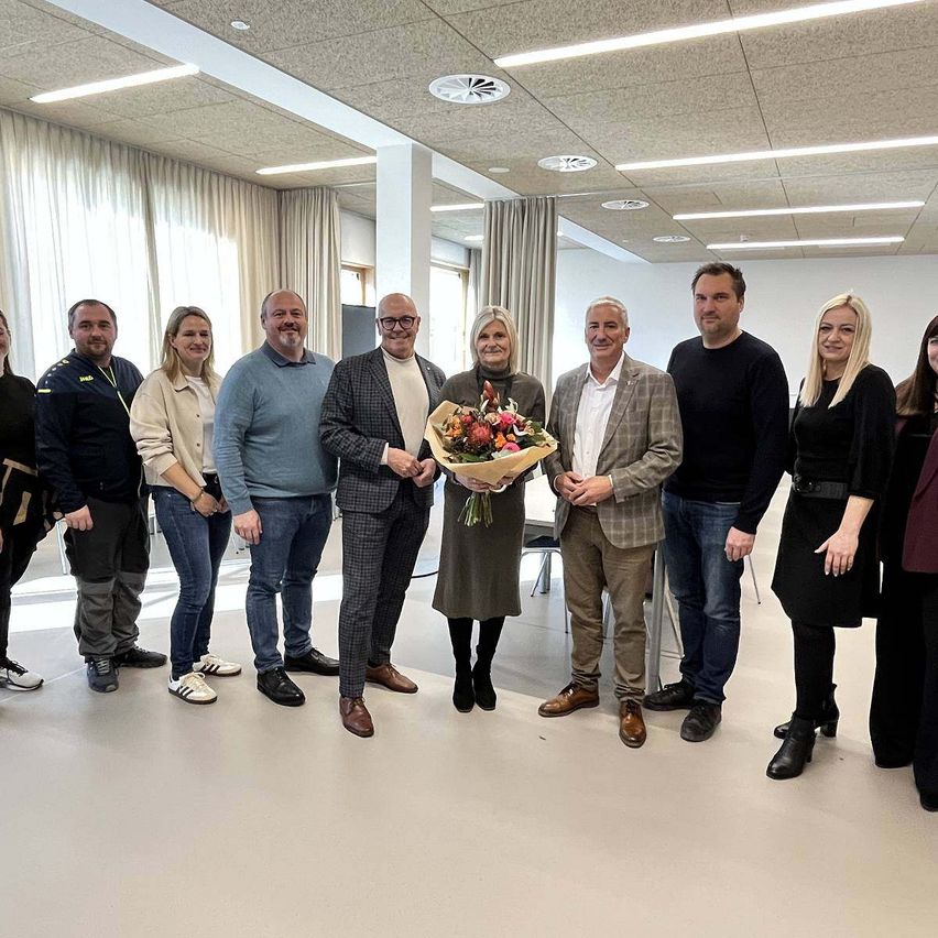 Eine Gruppe von Menschen steht in einem Raum, zwei von ihnen halten einen Blumenstrauß. Sie sind in Freizeitkleidung gekleidet. Der Raum hat weiße Wände und Vorhänge und einen weißen Boden.