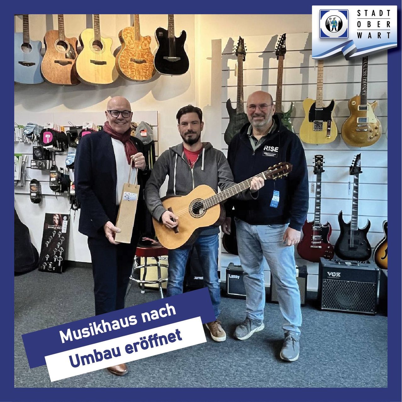 Drei Männer in einem Musikgeschäft posieren mit Gitarren im Hintergrund. Der Mann in der Mitte hält eine Gitarre. Der Text überlagert sagt 'Musikhaus nach Umbau eröffnet'.