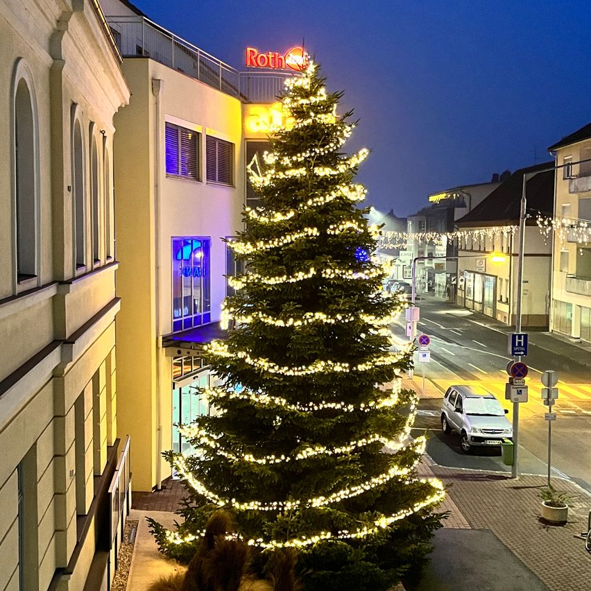 Ein großer Weihnachtsbaum mit Lichtern ist auf der Straßenecke vor einem Gebäude mit einem Roth-Schild oben dekoriert.