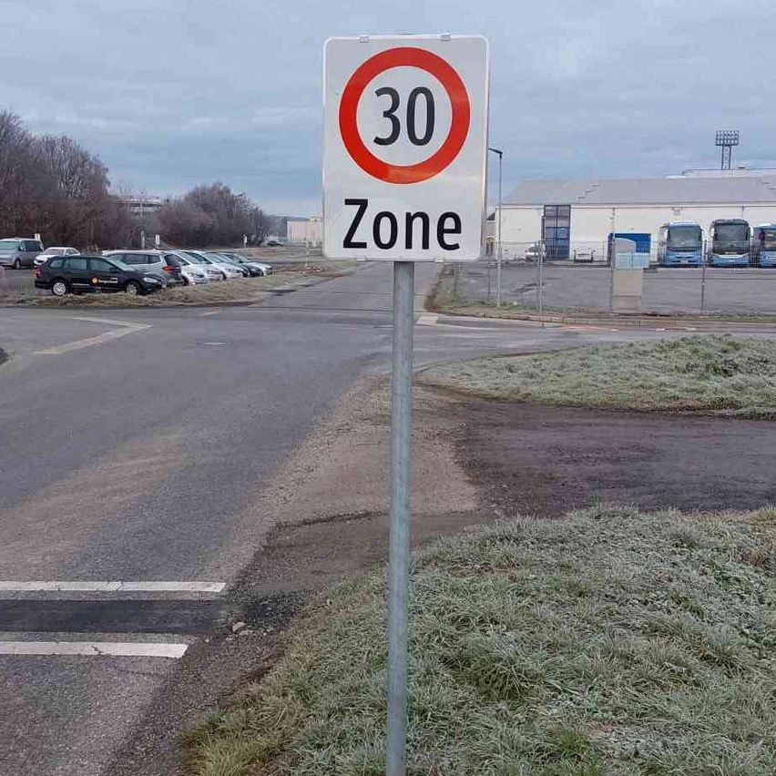 Ein Schild, das eine 30 km/h Geschwindigkeitsbegrenzung anzeigt, steht an einer Straße, mit einem Parkplatz und mehreren geparkten Autos im Hintergrund.