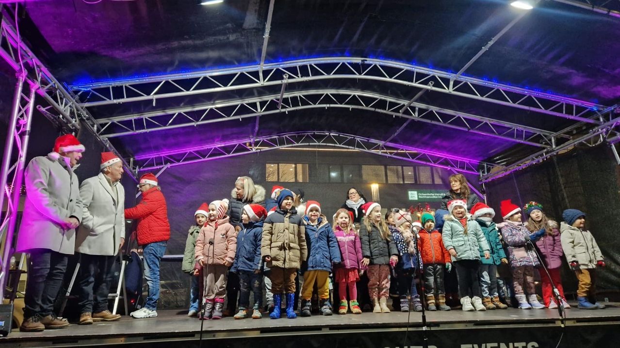 Eine Gruppe von Kindern in Winterkleidung und Weihnachtsmützen steht auf einer Bühne mit Erwachsenen unter einer Struktur mit lila und blauen Lichtern.
