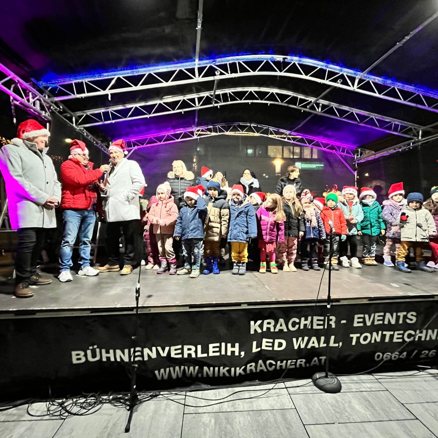 Eine Gruppe von Kindern und Erwachsenen steht auf einer Bühne, einige tragen Weihnachtsmützen. Sie scheinen an einer Weihnachtsveranstaltung teilzunehmen.